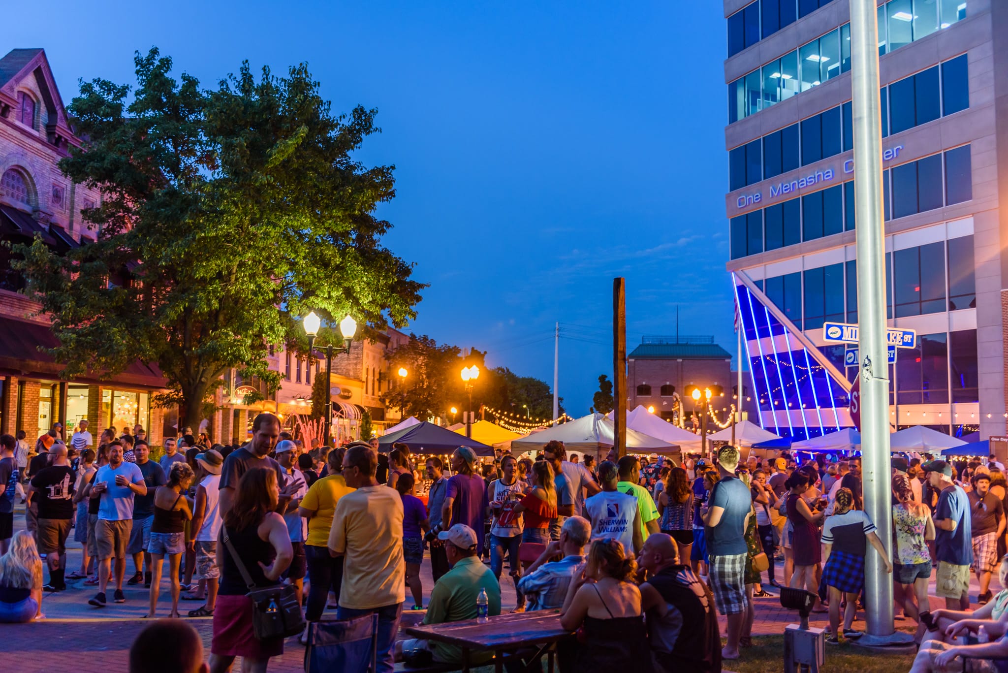 Crowd at an outdoor festival at dusk with tents, string lights, and a modern building.