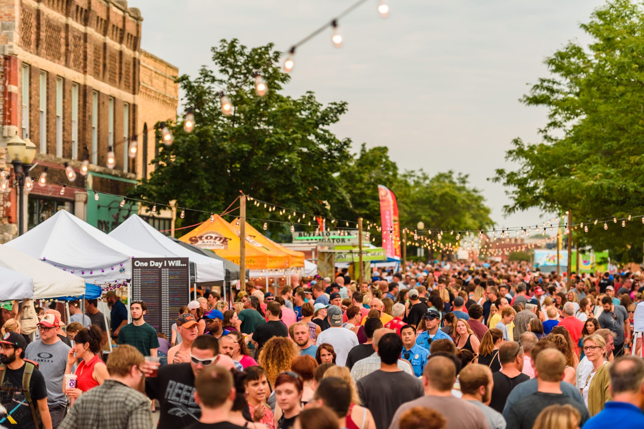 Crowd at outdoor festival with tents, food vendors, and string lights.
