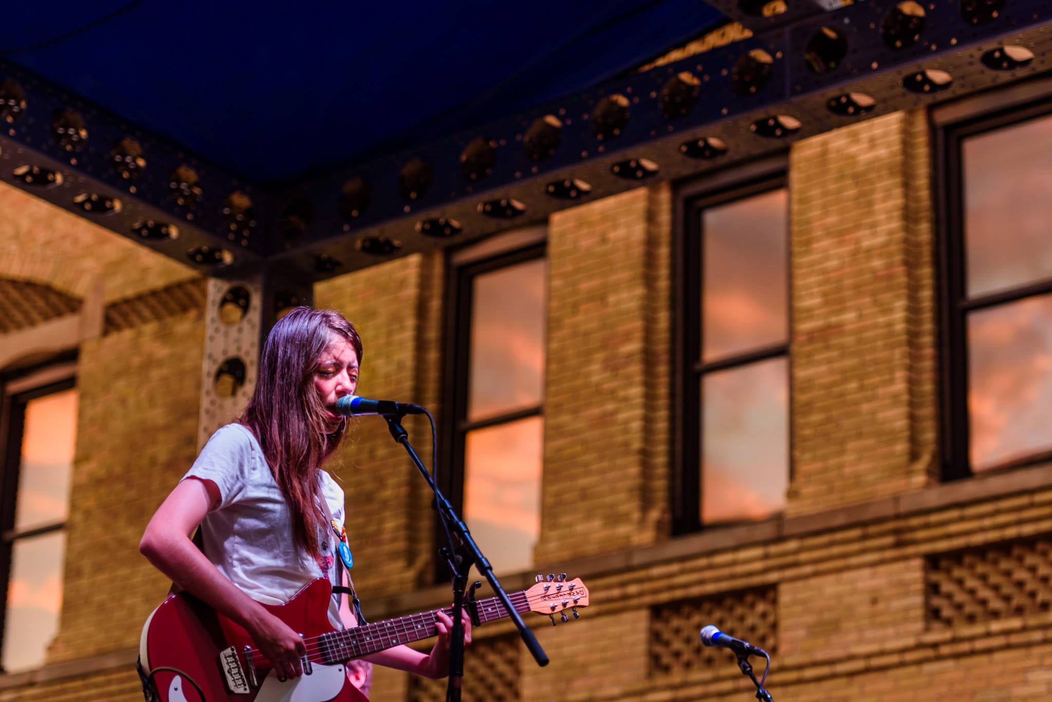 Woman singing and playing a red electric guitar on stage with a brick building background.