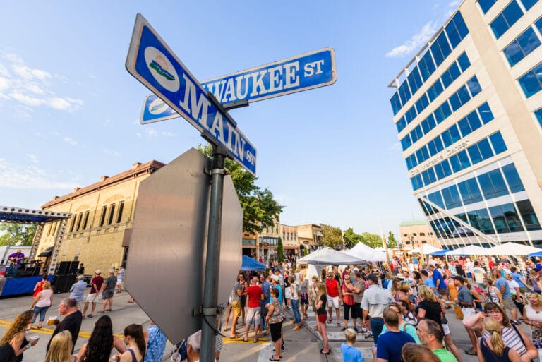 Street signs for Milwaukee St and Main St at a crowded outdoor festival with tents and buildings.