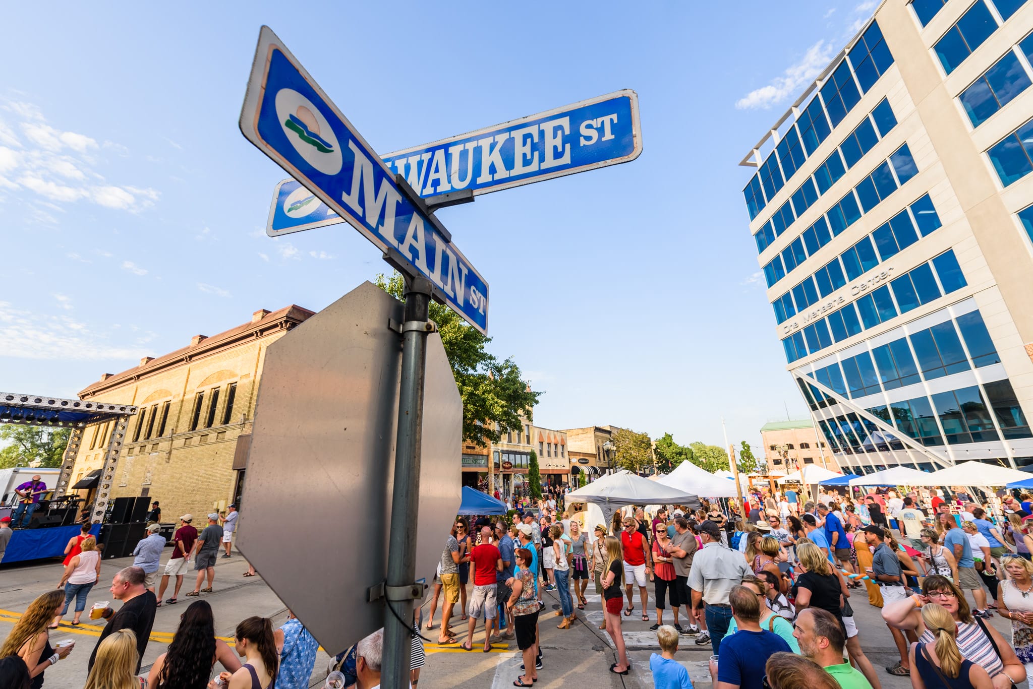 Street signs for Milwaukee St and Main St at a crowded outdoor festival with tents and buildings.