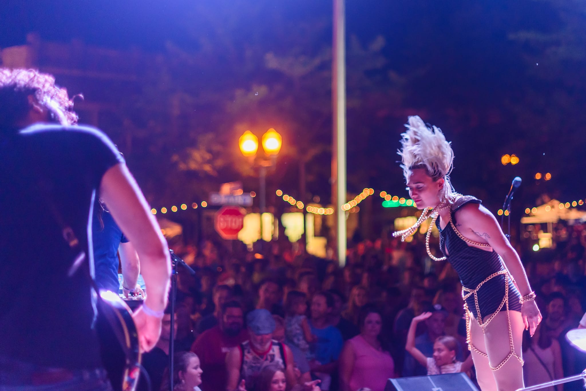 Female performer with spiked hair in a black outfit with gold chains, on stage before a crowd.