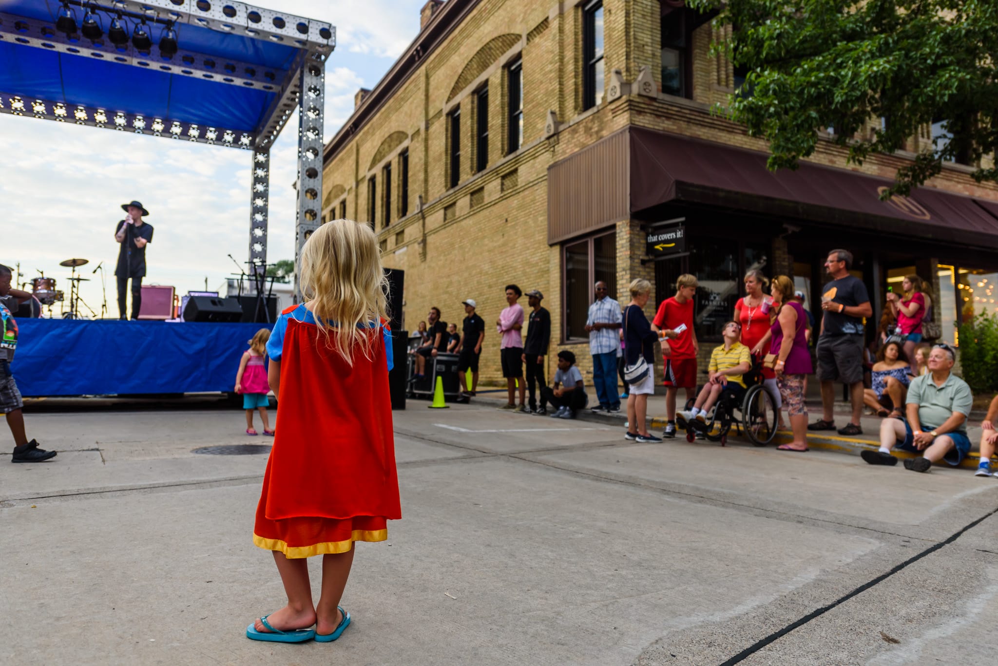 Girl in superhero cape watches performer on stage at outdoor event.