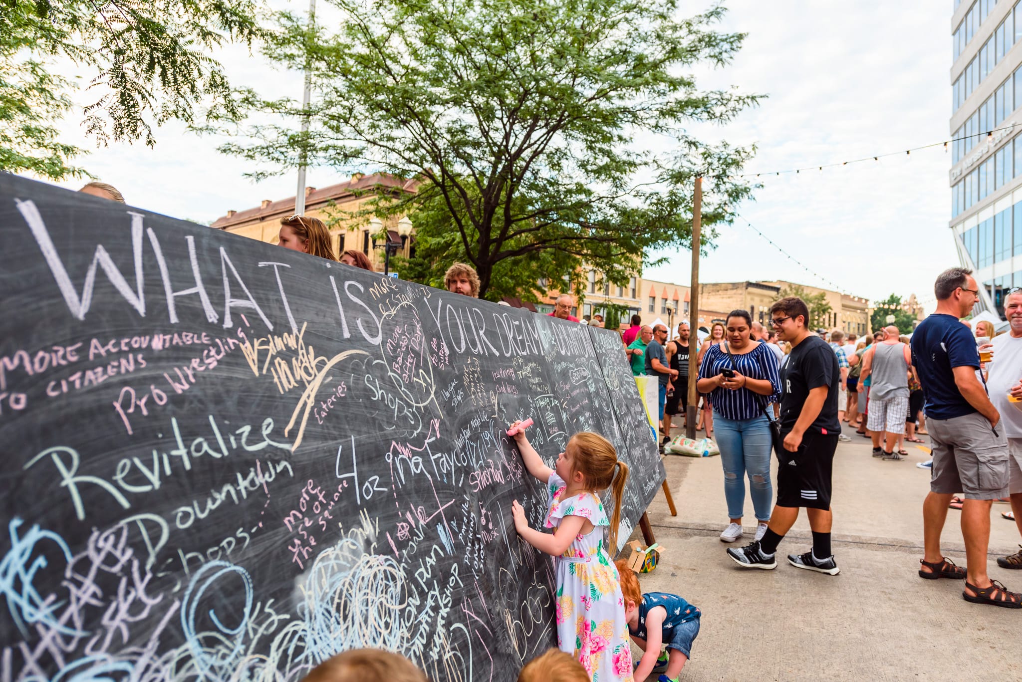 Children and adults write on a large chalkboard with chalk messages.