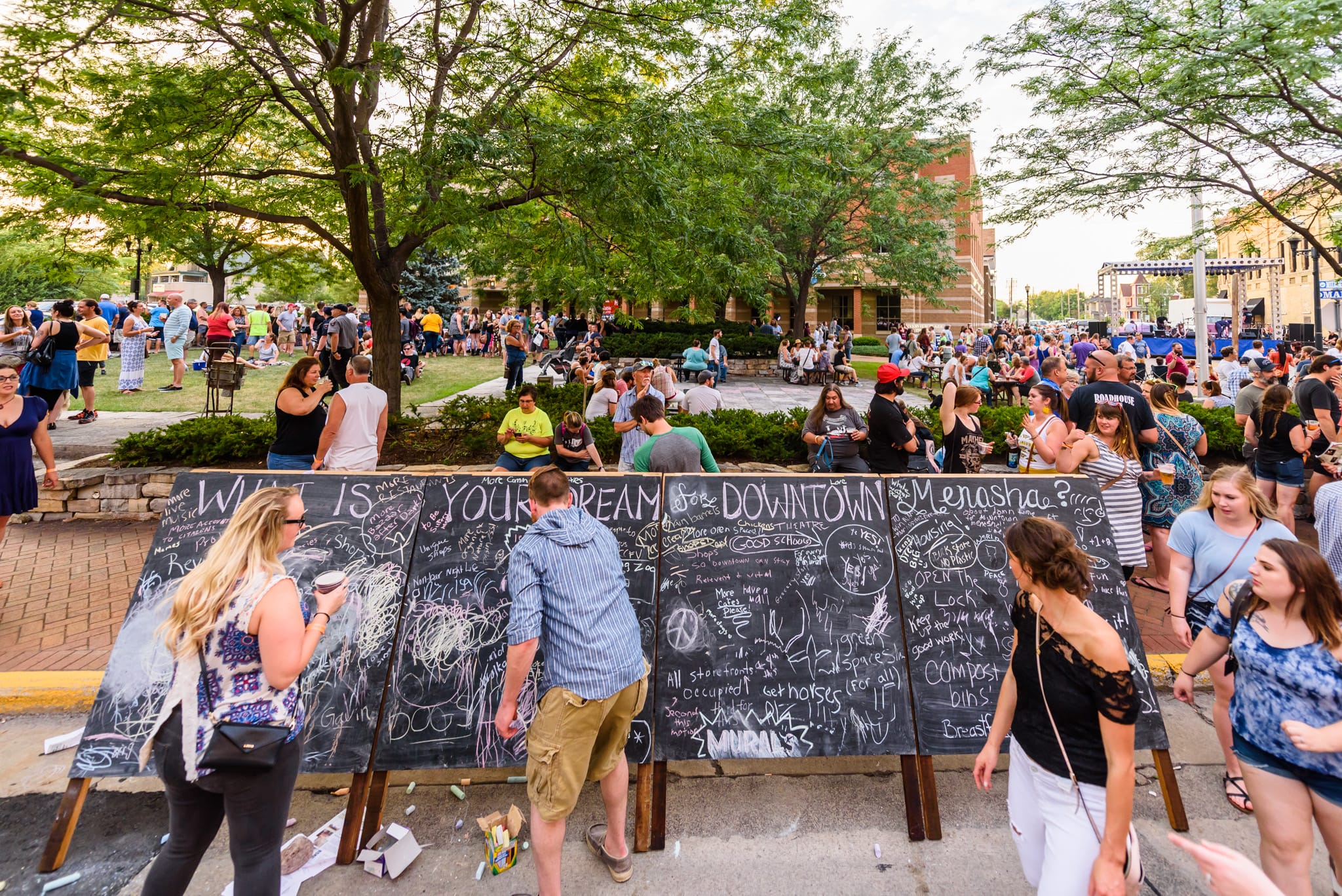 People write on chalkboards at a street festival with a crowd in the background.