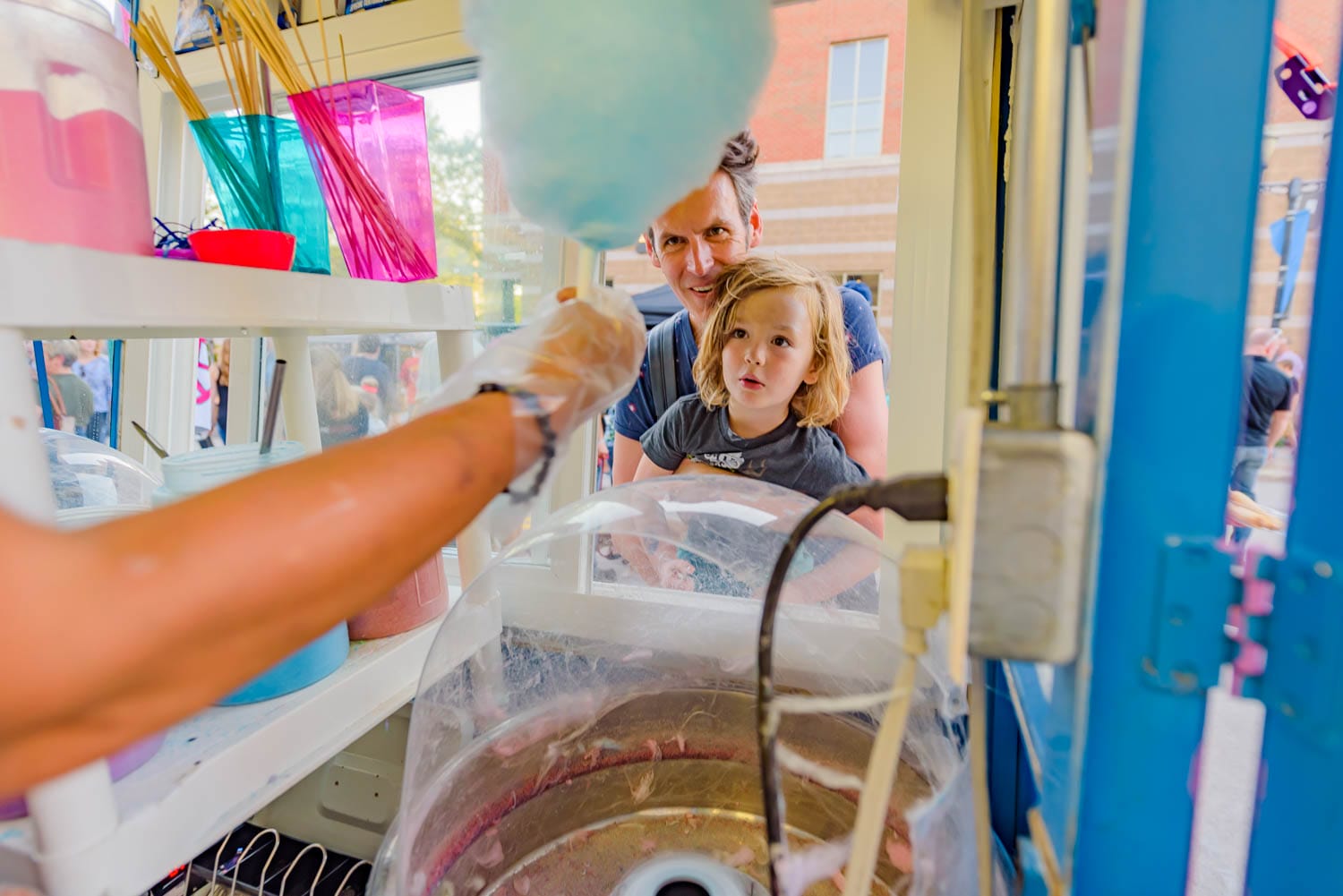 Father and child watch blue cotton candy being made at a stand.