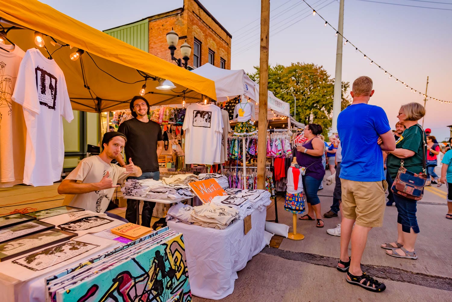 Vendors at an outdoor market display t-shirts and art under a yellow tent.