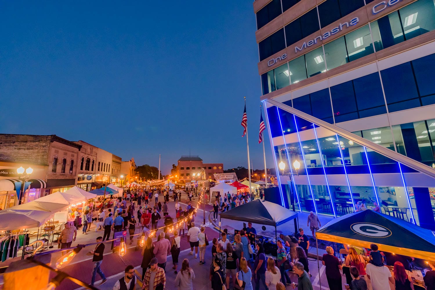 Street festival at dusk with tents, crowds, and a modern building with blue lights.