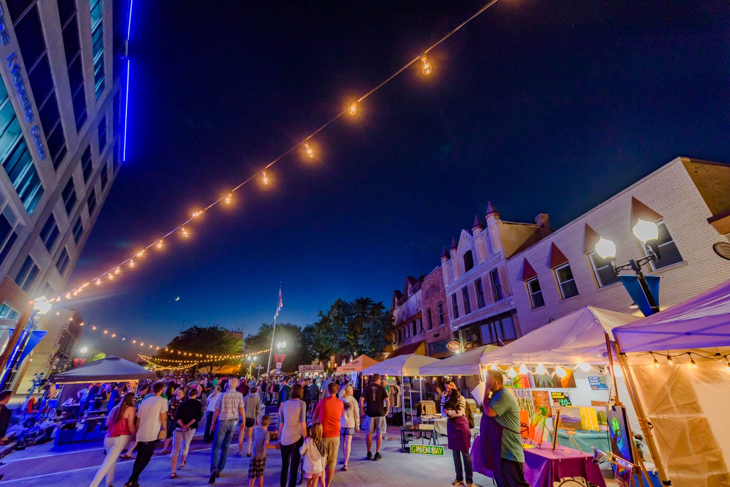 Street fair at night with string lights, tents, and people browsing.