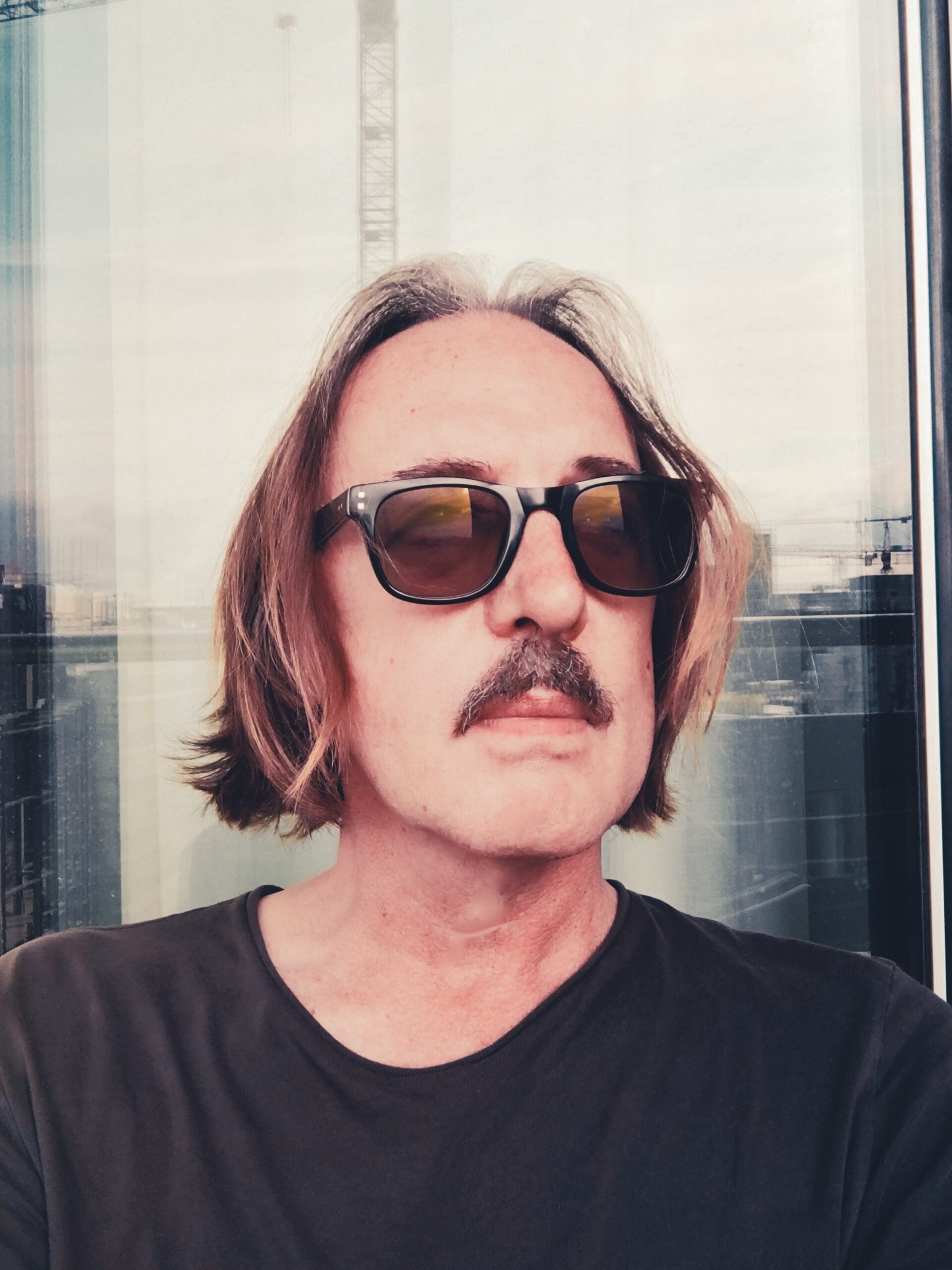 Man with mustache and sunglasses, reflected in a window with a crane in the background.