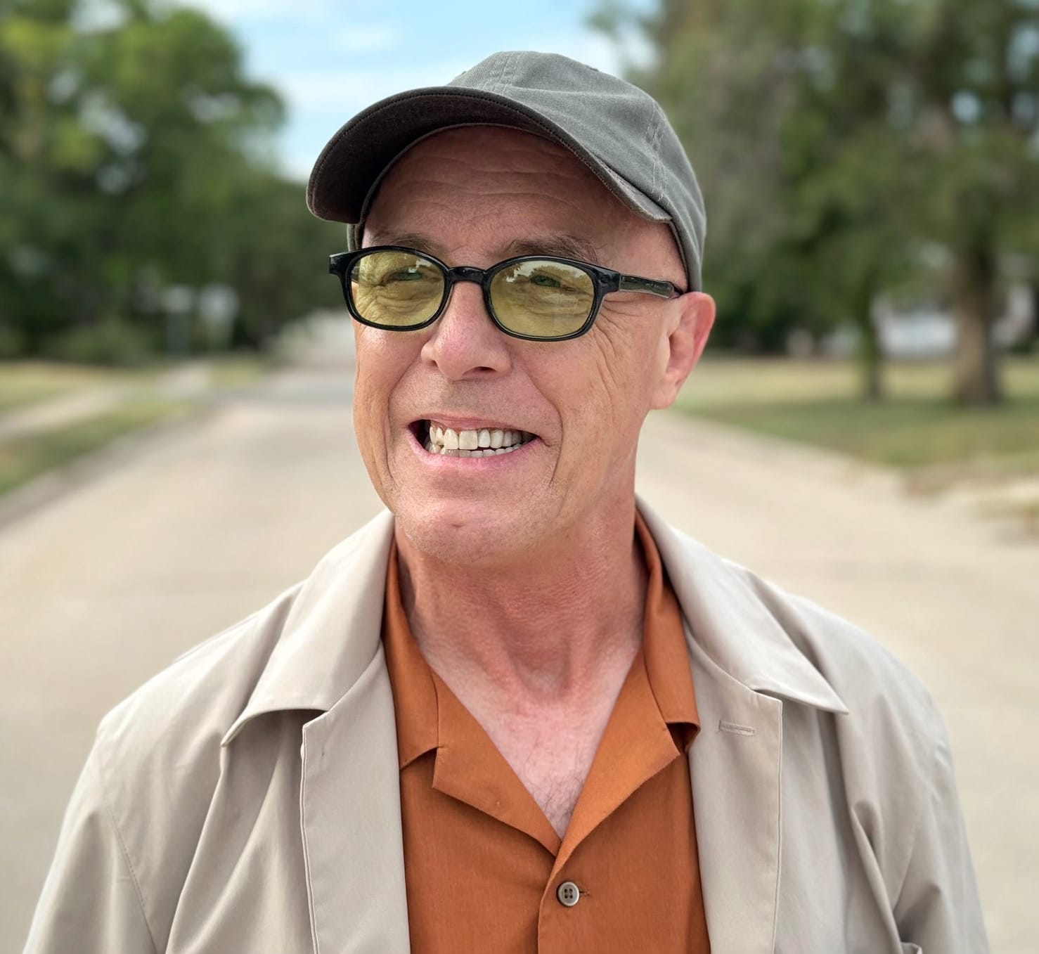 Man in cap and glasses smiles, wearing a tan jacket over an orange shirt.