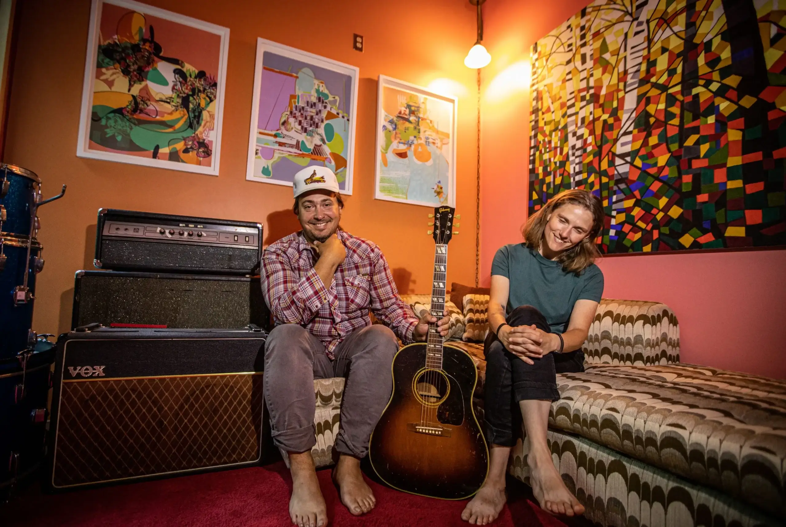 Two musicians sit with a guitar and amps, colorful art on the wall.