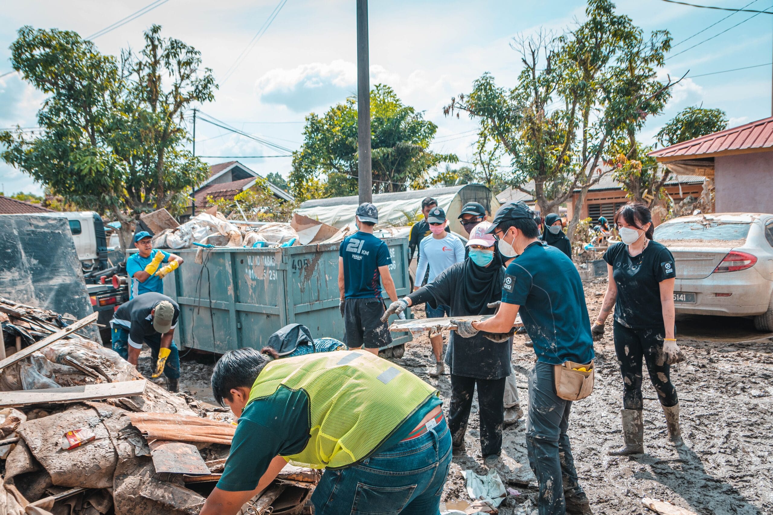 Volunteers in masks and gloves clear debris after a flood, with a dumpster and damaged car visible.