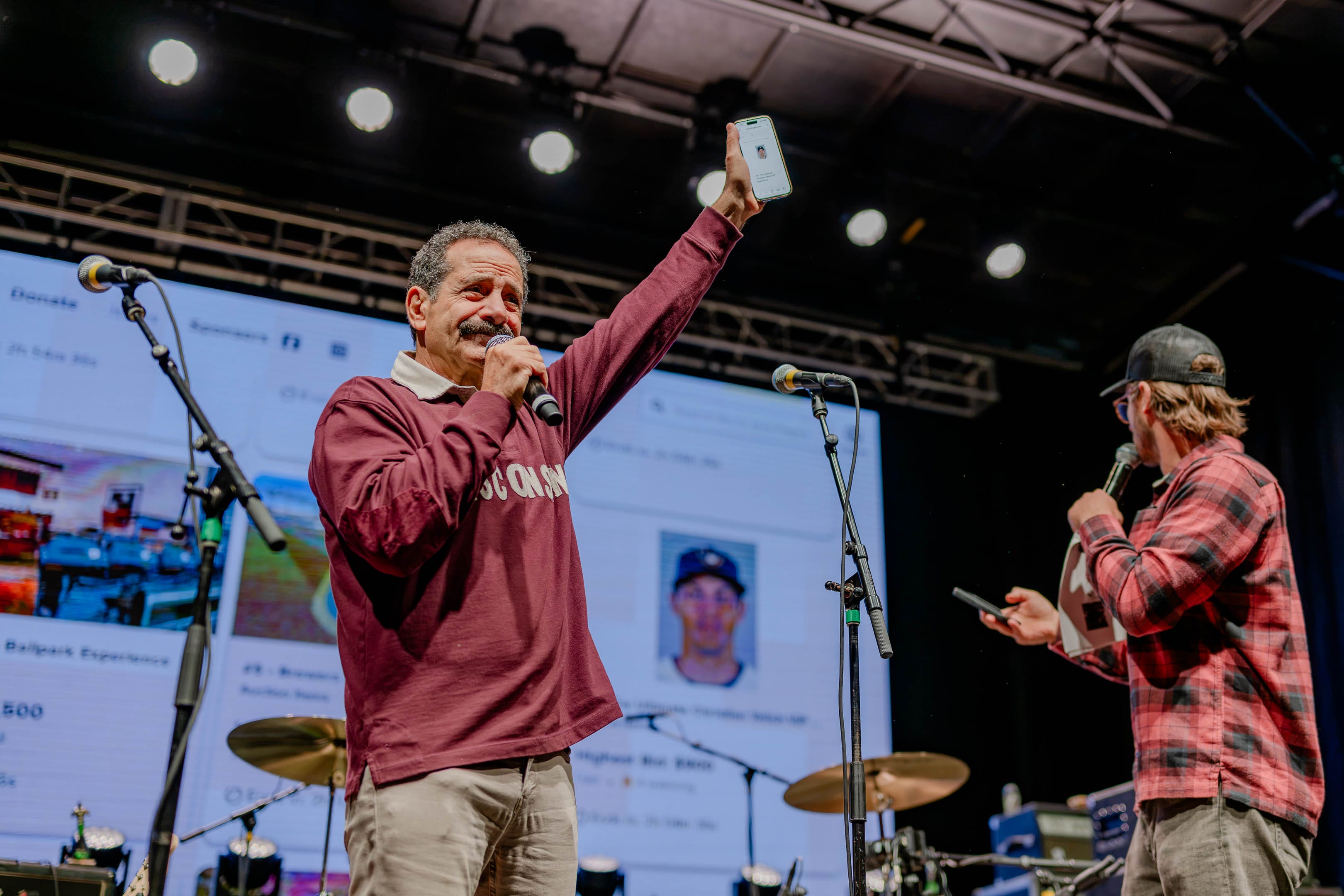 Man in maroon sweatshirt holds up phone and microphone on stage with a large screen behind him.